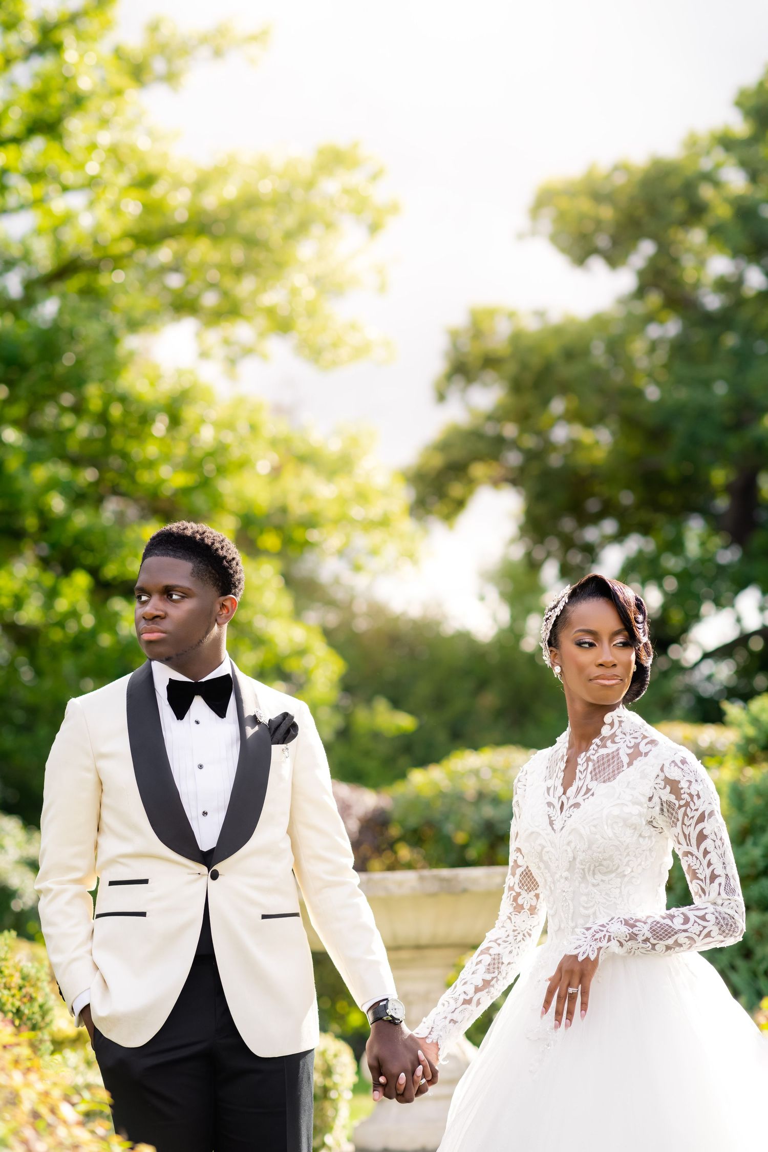 Newlyweds pose together in a garden setting with sunlit trees in the background.