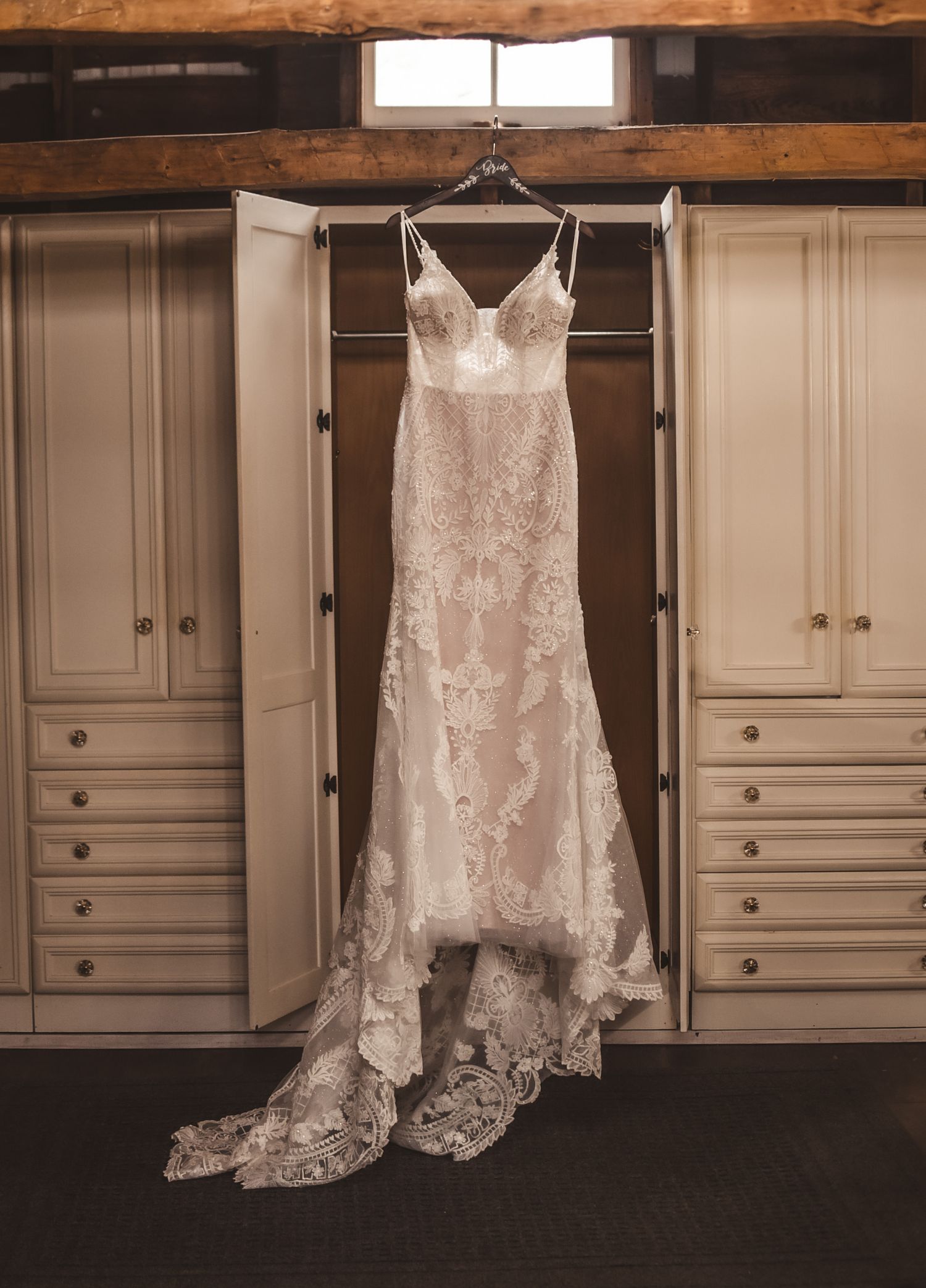 Elegant white lace wedding dress with sweetheart neckline hanging on display between cream-colored built-in cabinets.