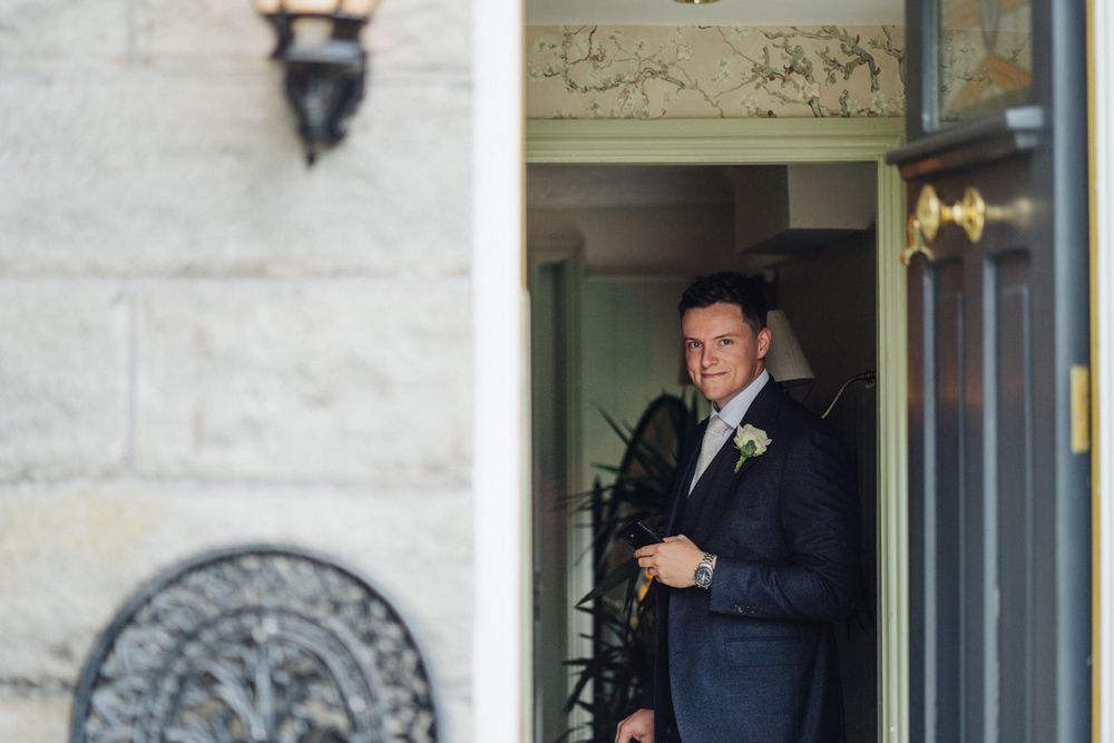 A groom in a navy suit stands in a doorway with ornate architectural details.