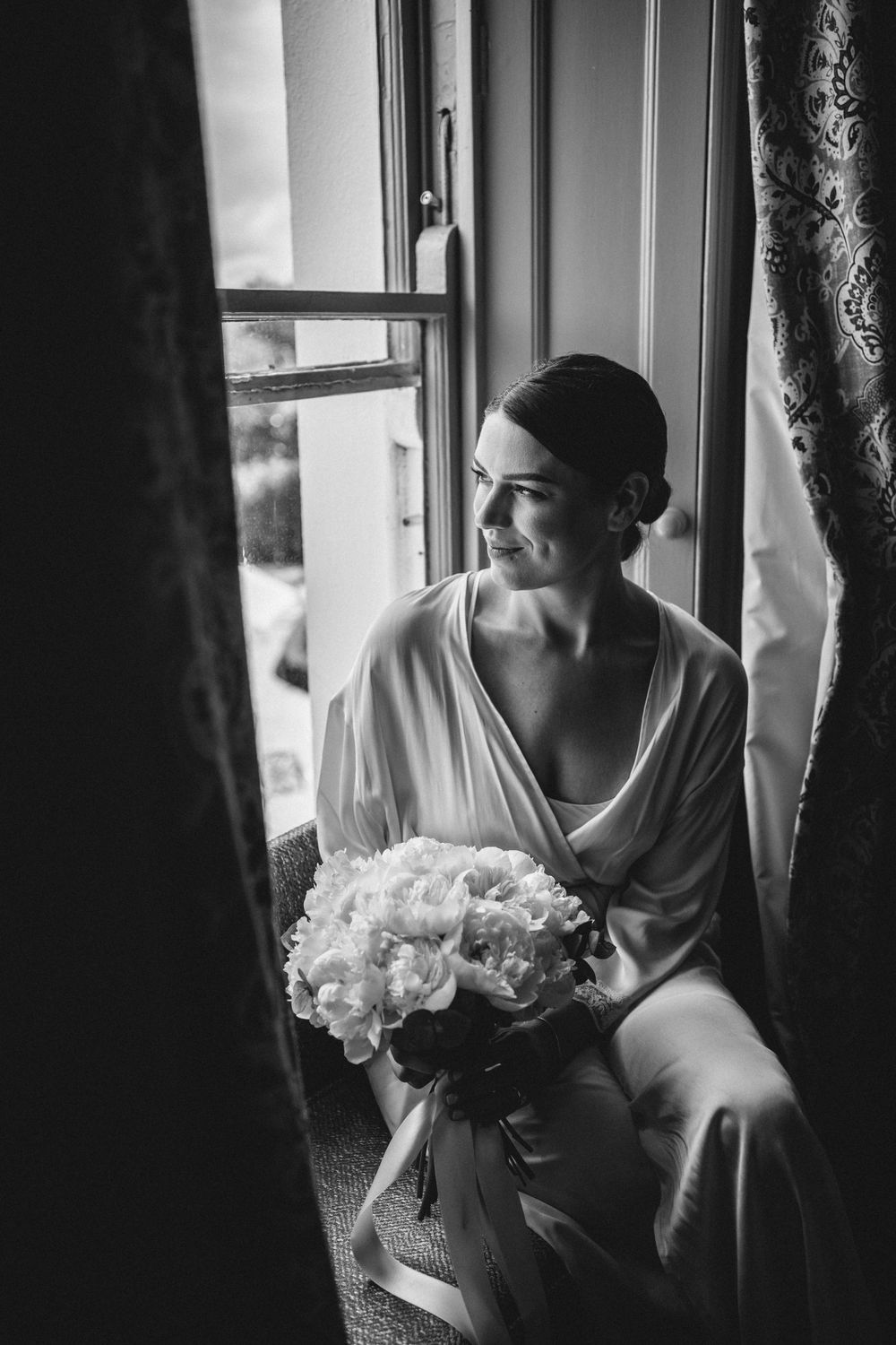 A person in a white robe holds a bouquet of white flowers while sitting by a window.