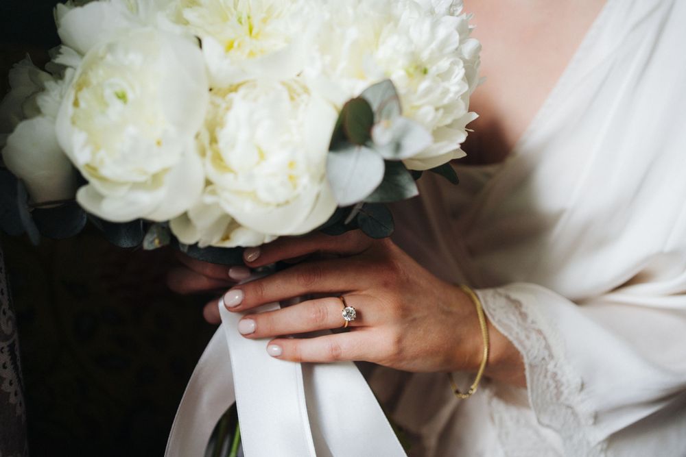 White peonies in a bridal bouquet with hands holding ribbon and wearing an engagement ring.