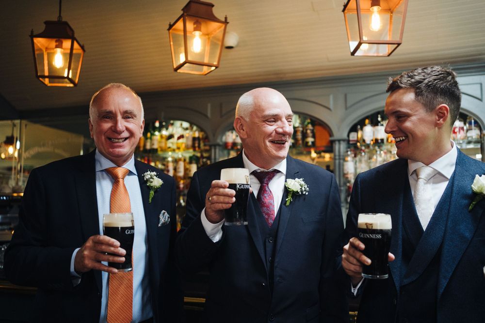 Groomsmen laughing and enjoying drinks at bar with vintage lantern lighting.
