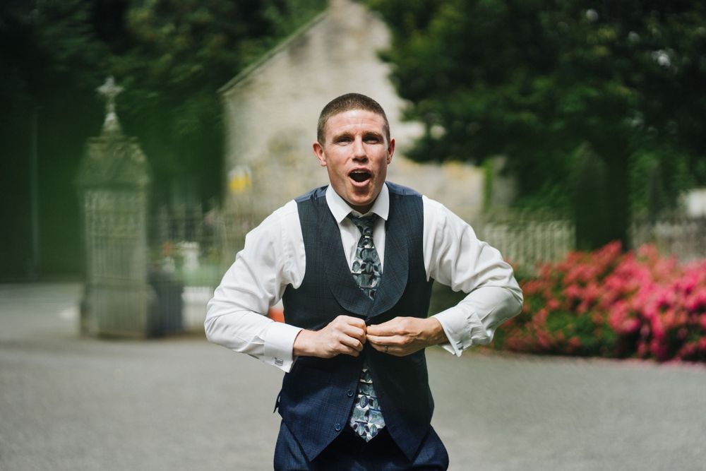 Person in formal vest and tie making animated expression outdoors by garden.