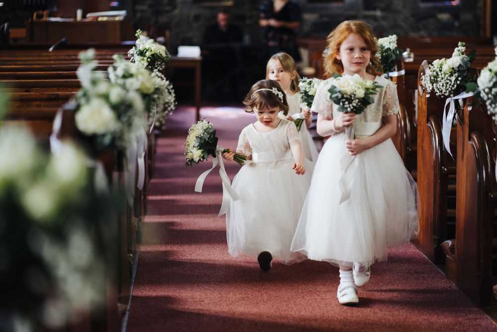 Flower girls in white dresses walking down church aisle decorated with flowers.