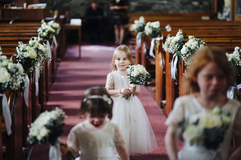 Small flower girls carrying bouquets down decorated church aisle.