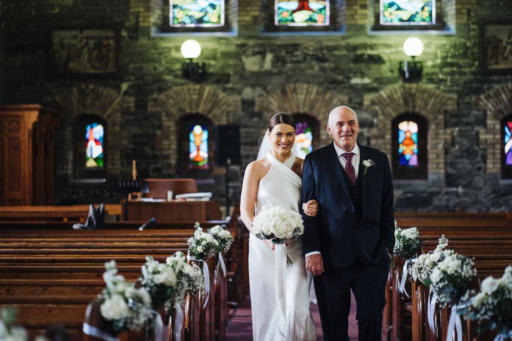 Wedding ceremony in a stone church with stained glass windows and floral-decorated pews along the aisle.