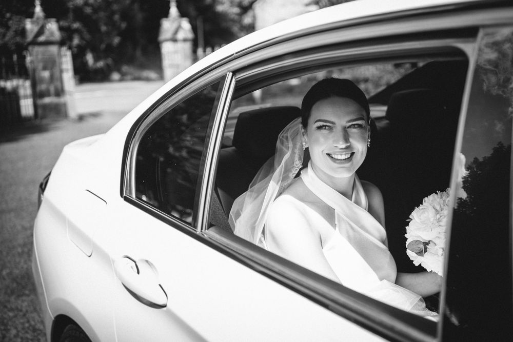 Black and white portrait of someone smiling through a car window with a wedding bouquet.