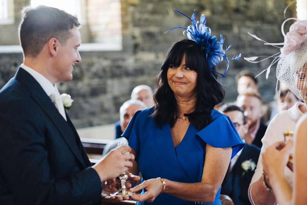 Wedding guest in vibrant blue fascinator hat conversing during outdoor reception.