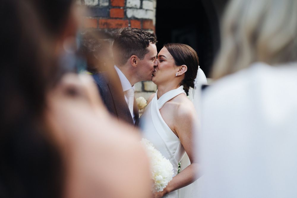 An intimate wedding kiss captured through a crowd of guests in a church setting.