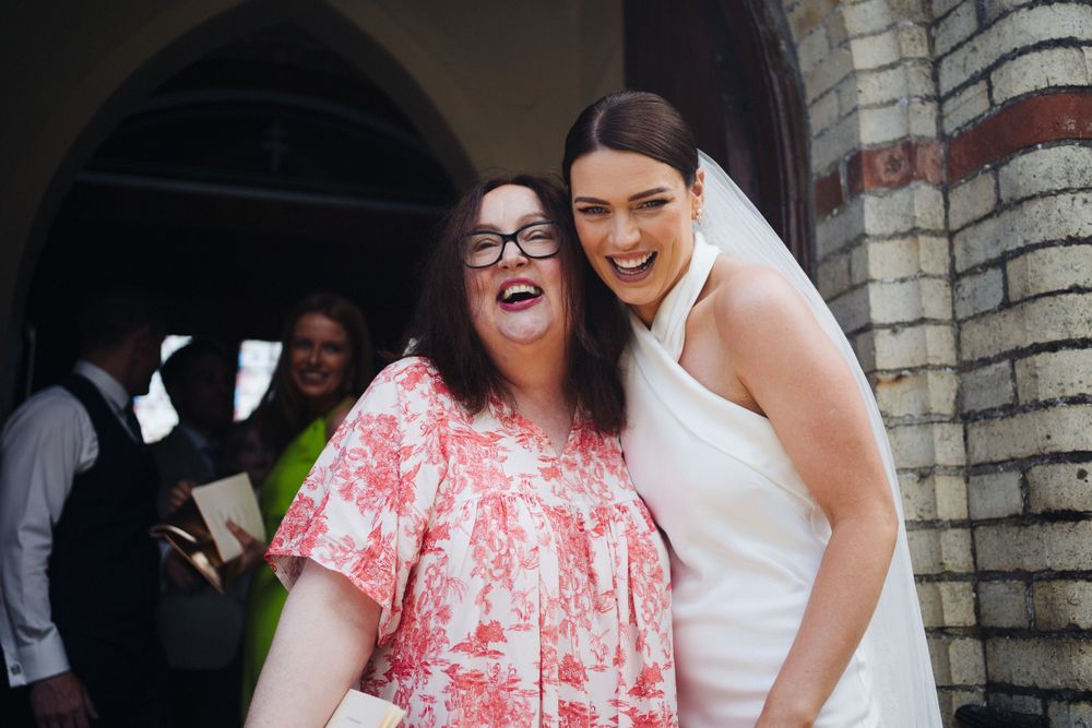 Two guests share an enthusiastic laugh under an arched doorway at a wedding.