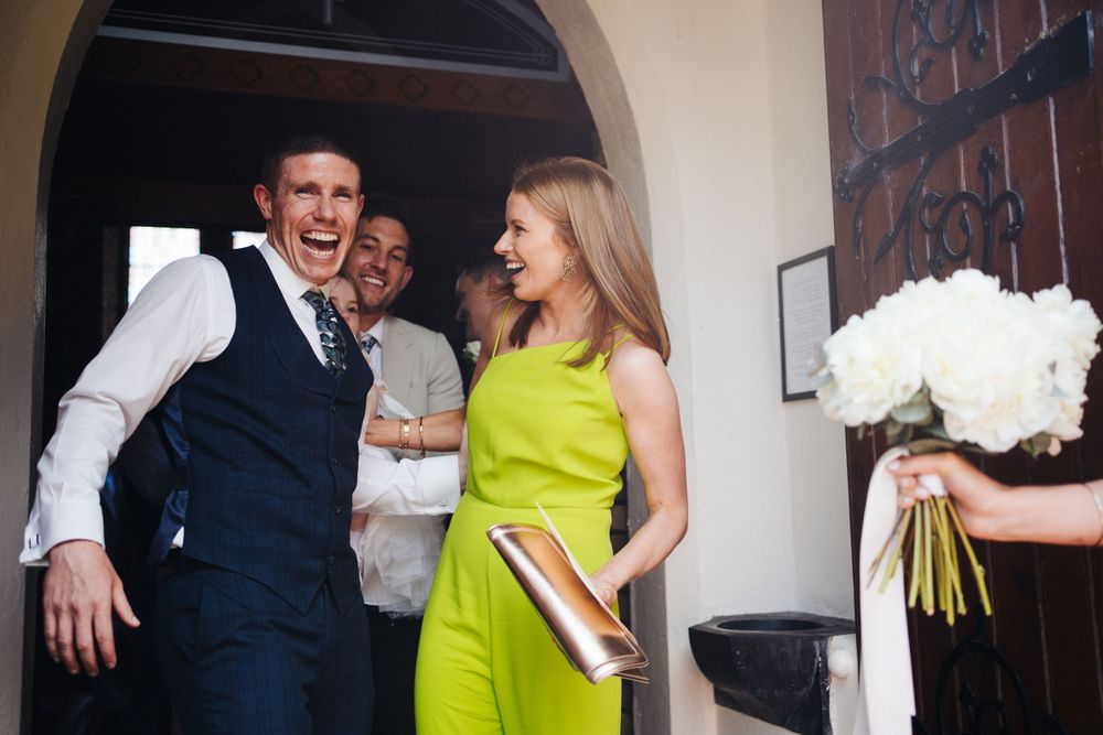 Wedding party members laugh joyfully at a reception entrance while someone holds a white bouquet.