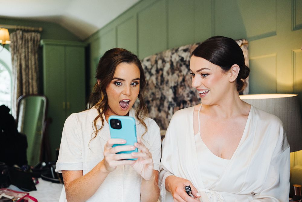 Two people in white robes share a lighthearted moment while looking at a phone in a green-walled room.