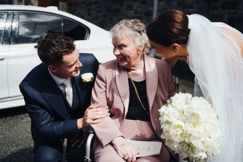 Wedding party members share a tender moment with elderly guest in pink outfit.