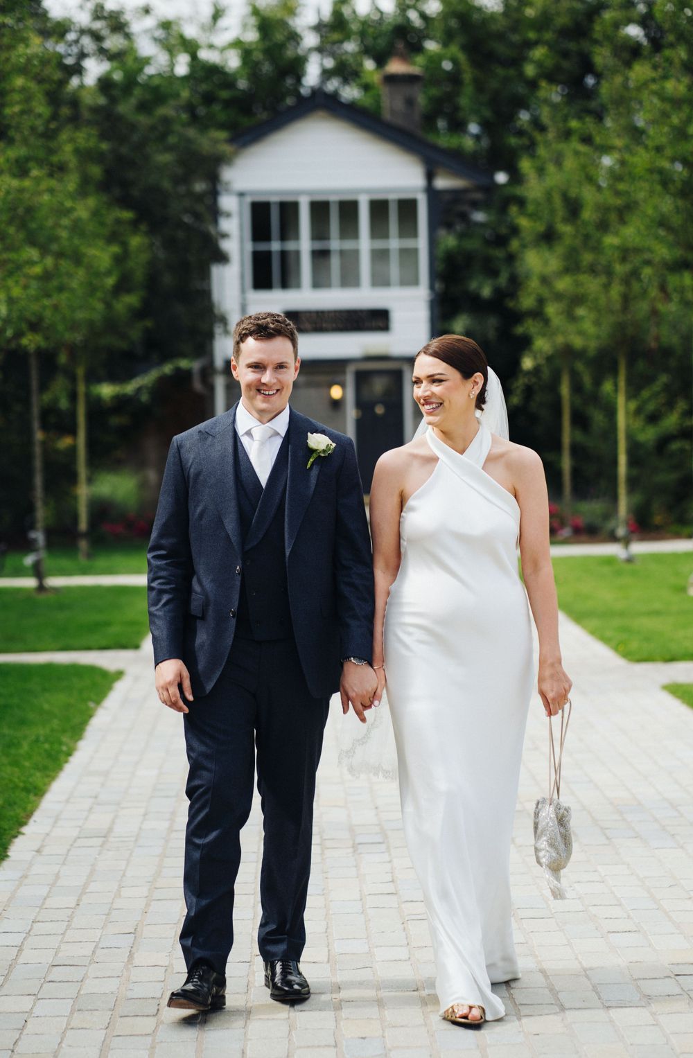 Couple walks together on stone path with white house and greenery in background.