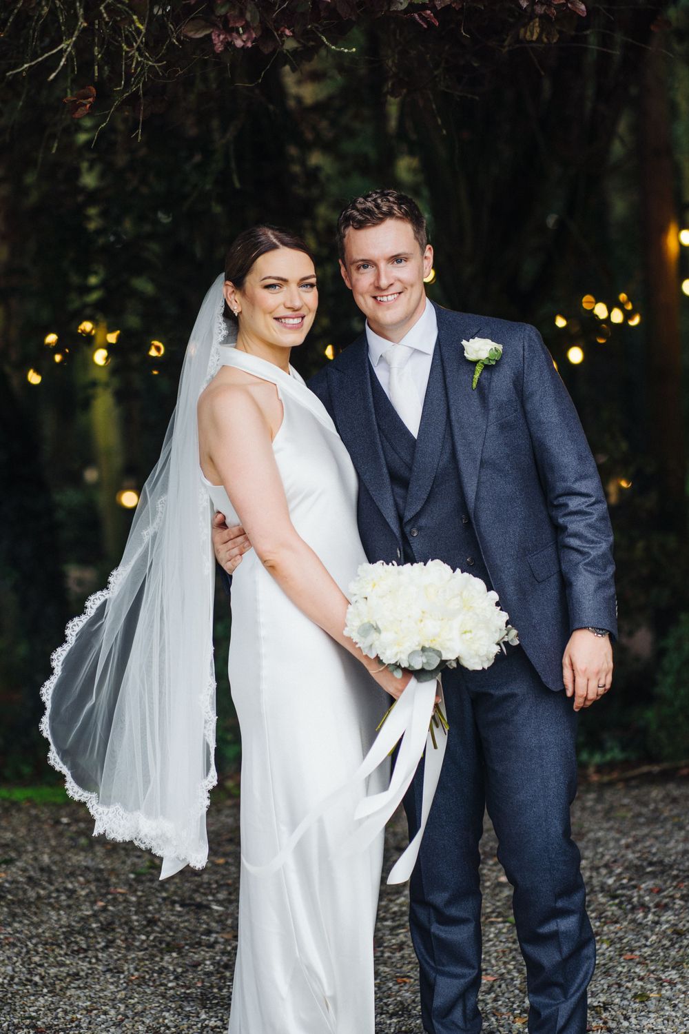 Evening wedding portrait with couple against backdrop of twinkle lights and greenery.