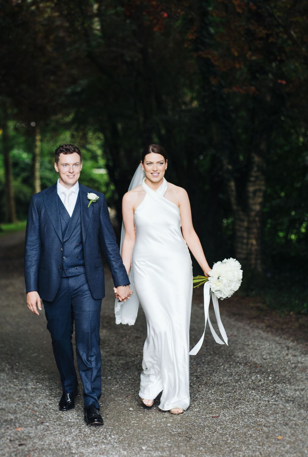 Newlyweds walk hand in hand on a garden path with the bride holding a white bouquet.