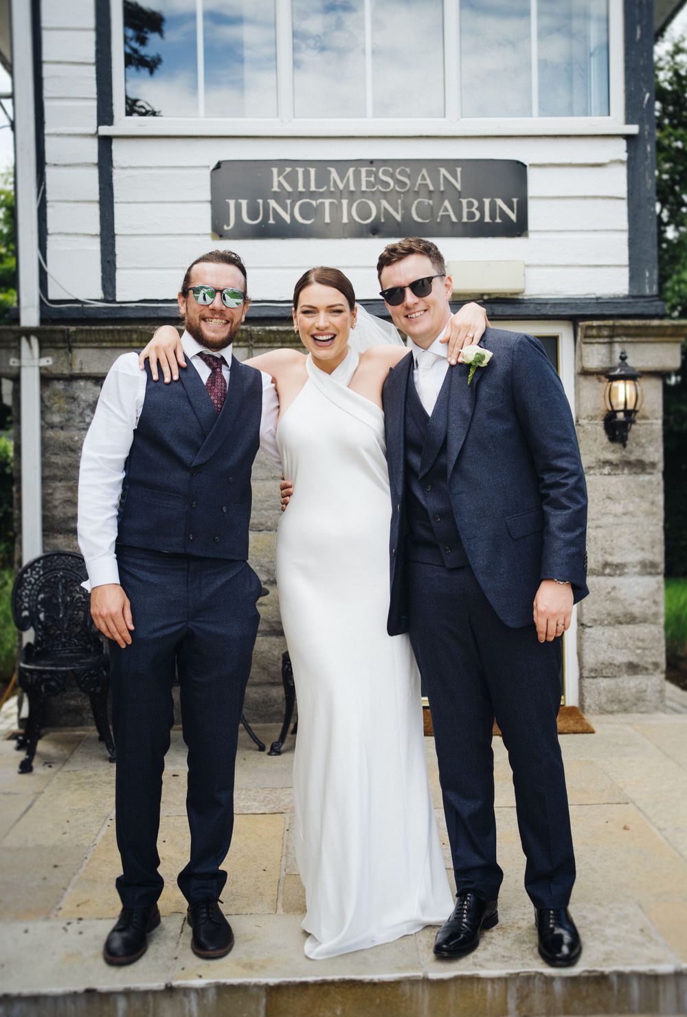 Wedding party poses outside Kilmessan Junction Cabin in formal attire.