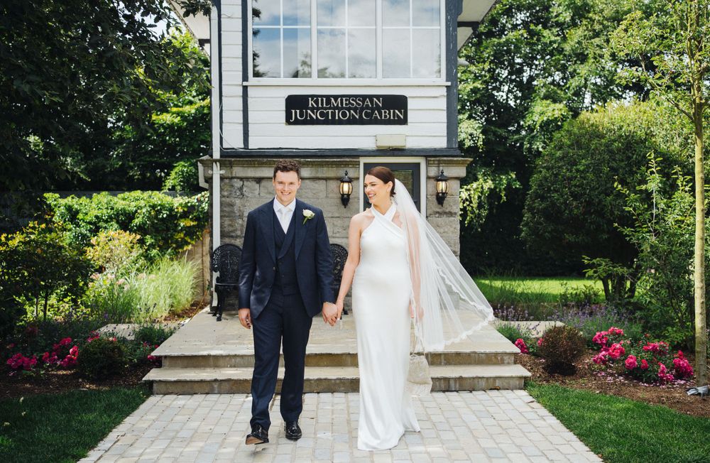 Elegant couple in wedding attire standing outside a building entrance with landscaped gardens.