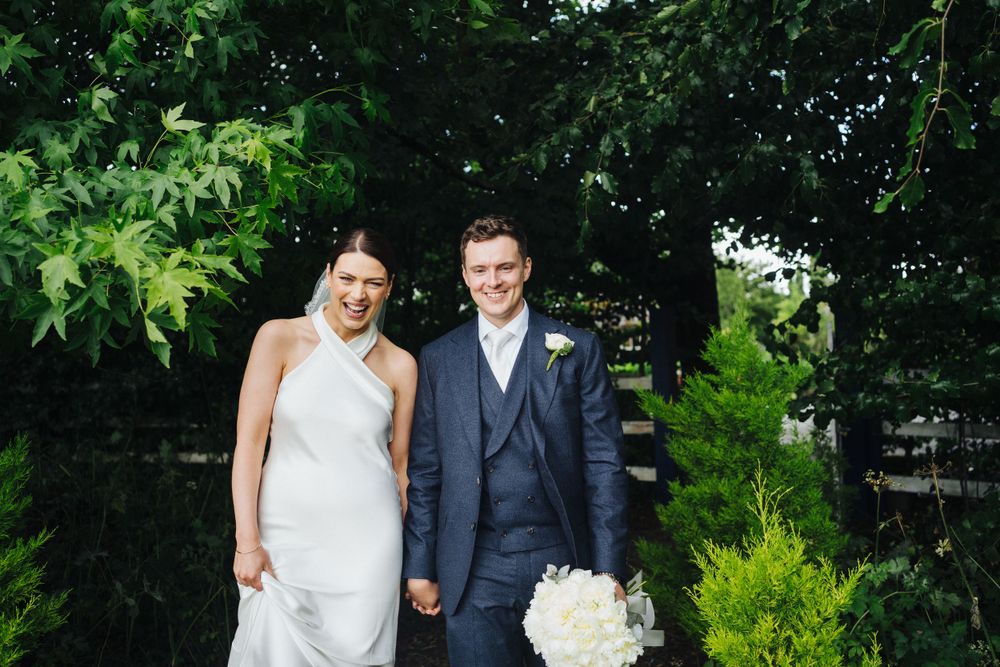 Wedding couple holding hands in a lush garden setting with white floral bouquet.