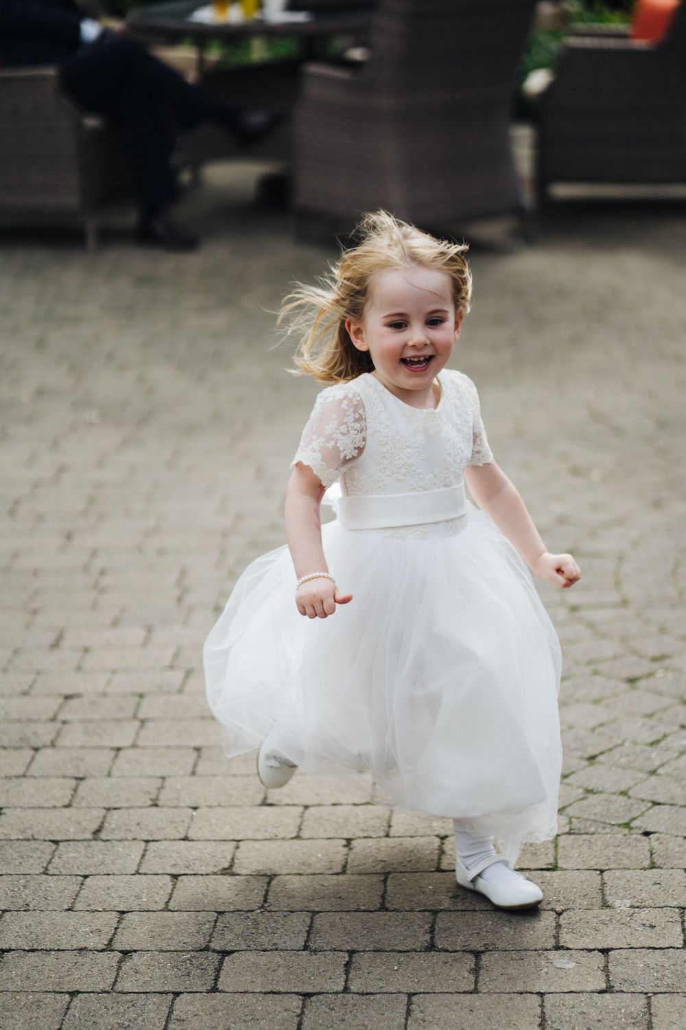 Young flower girl in white dress running joyfully on stone pavers.