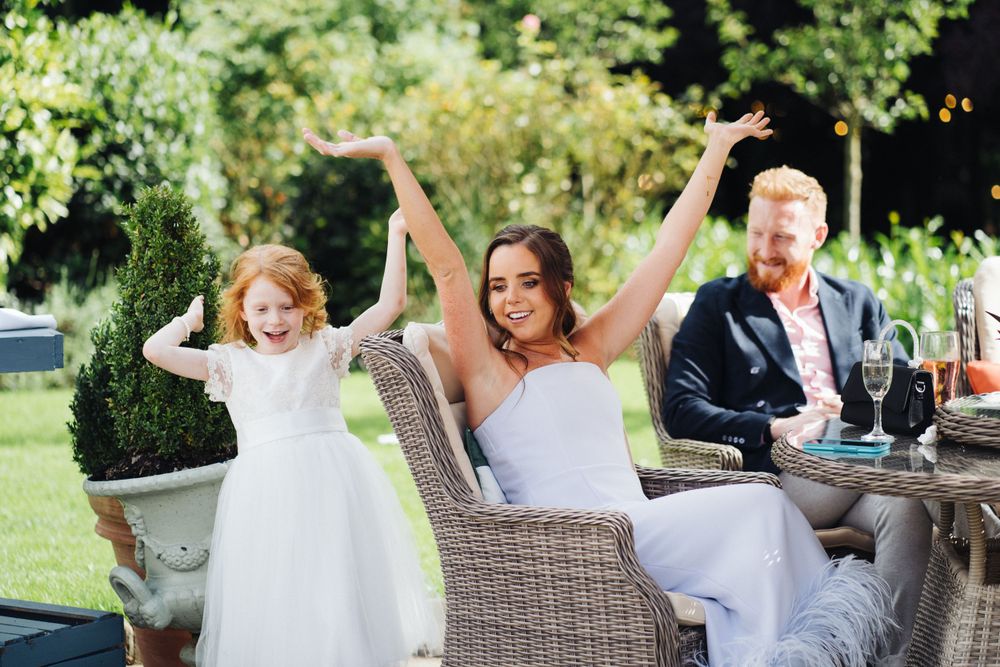 Wedding party celebrates joyfully at an outdoor garden reception with raised arms and excited expressions.