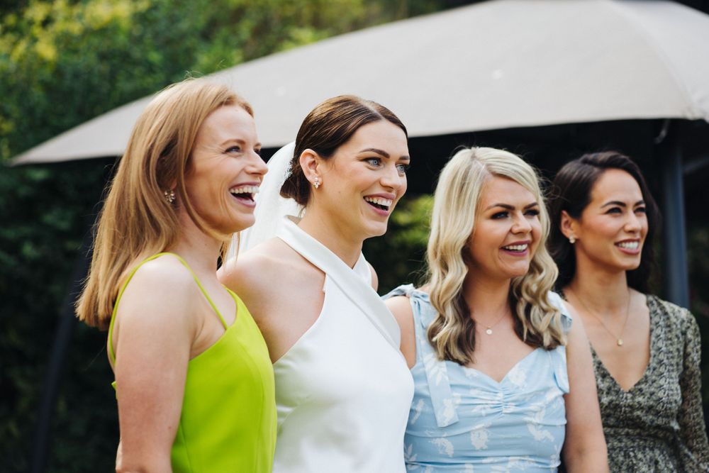 Four women in formal attire smile together at an outdoor celebration.