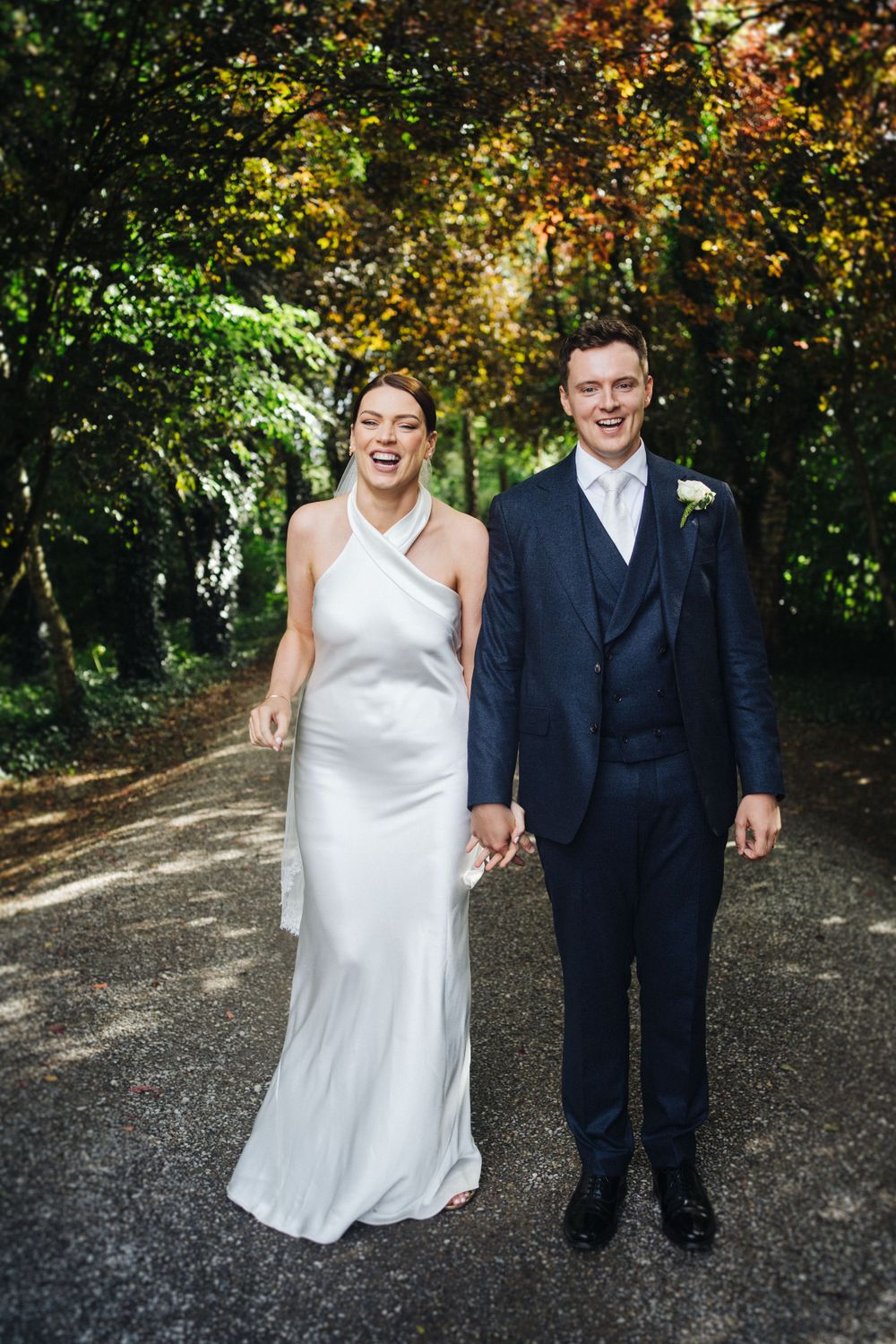 Happy couple in wedding attire laughing together on a wooded path.