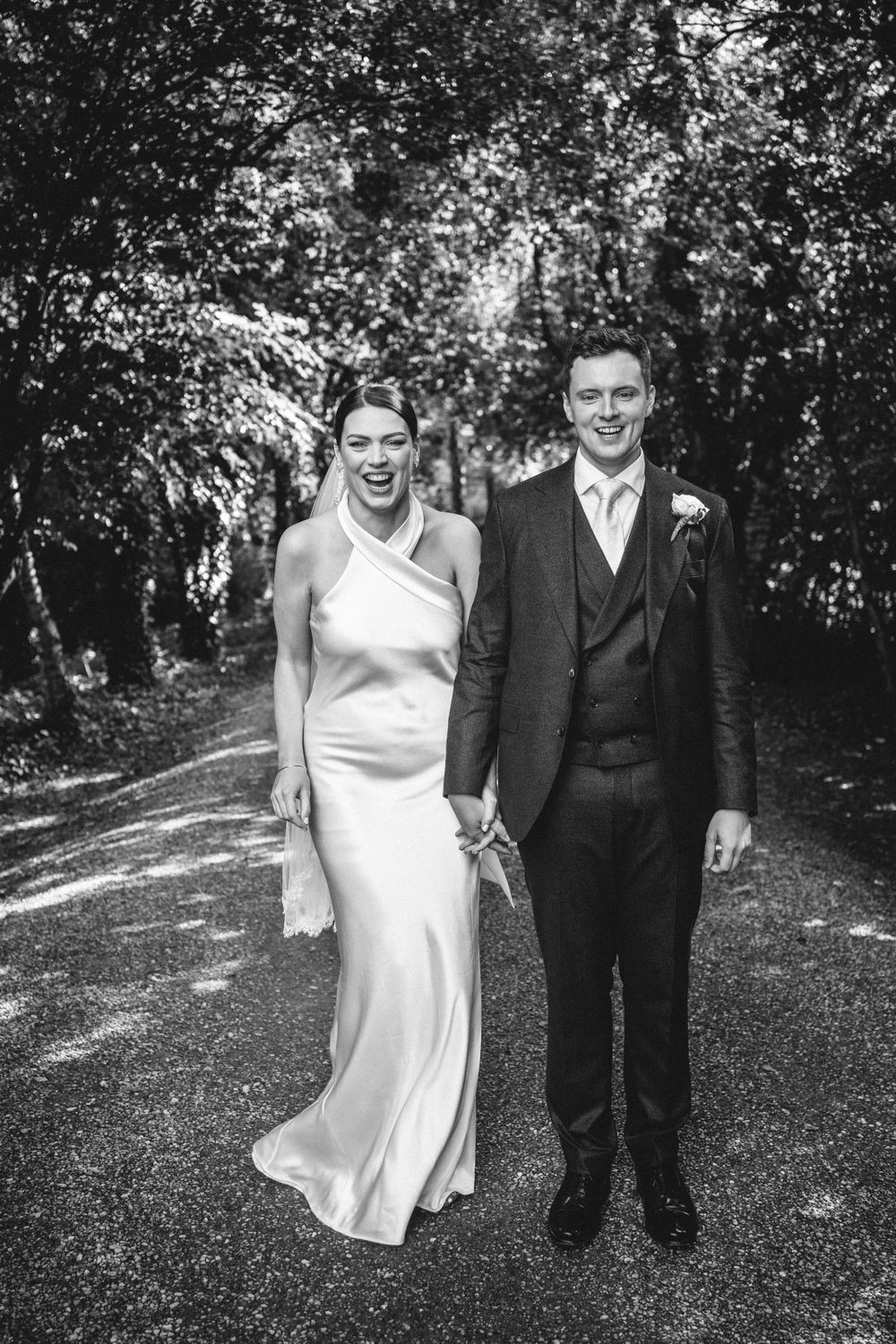 Black and white portrait of newlyweds standing on a tree-lined path.