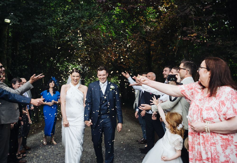 Wedding guests throw confetti over newlyweds walking down a forest path.