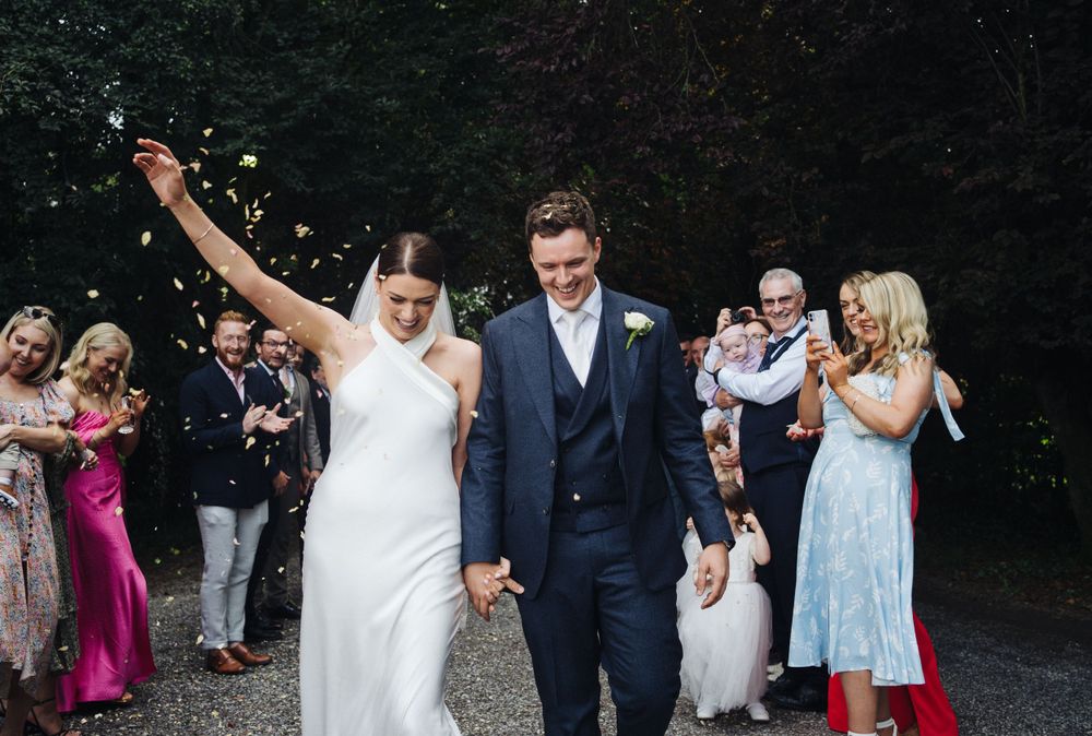 Bride raises arm triumphantly while walking through confetti with guests celebrating.