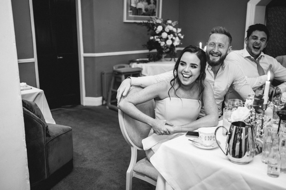 Candid black and white photo of reception guests laughing at dining table.