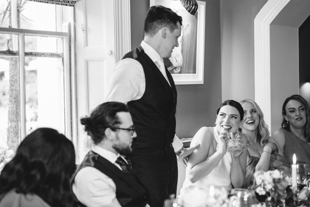 A wedding reception speaker stands and addresses guests at a decorated dining table.