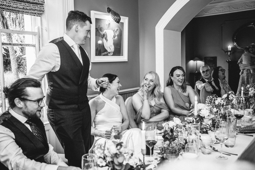 Wedding guests enjoy speeches at an elegant indoor reception with floral centerpieces.