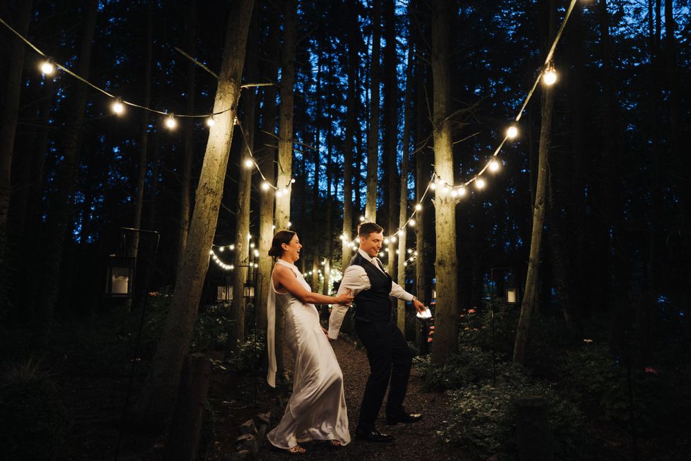 Couple dances under string lights in a romantic forest wedding setting at dusk.