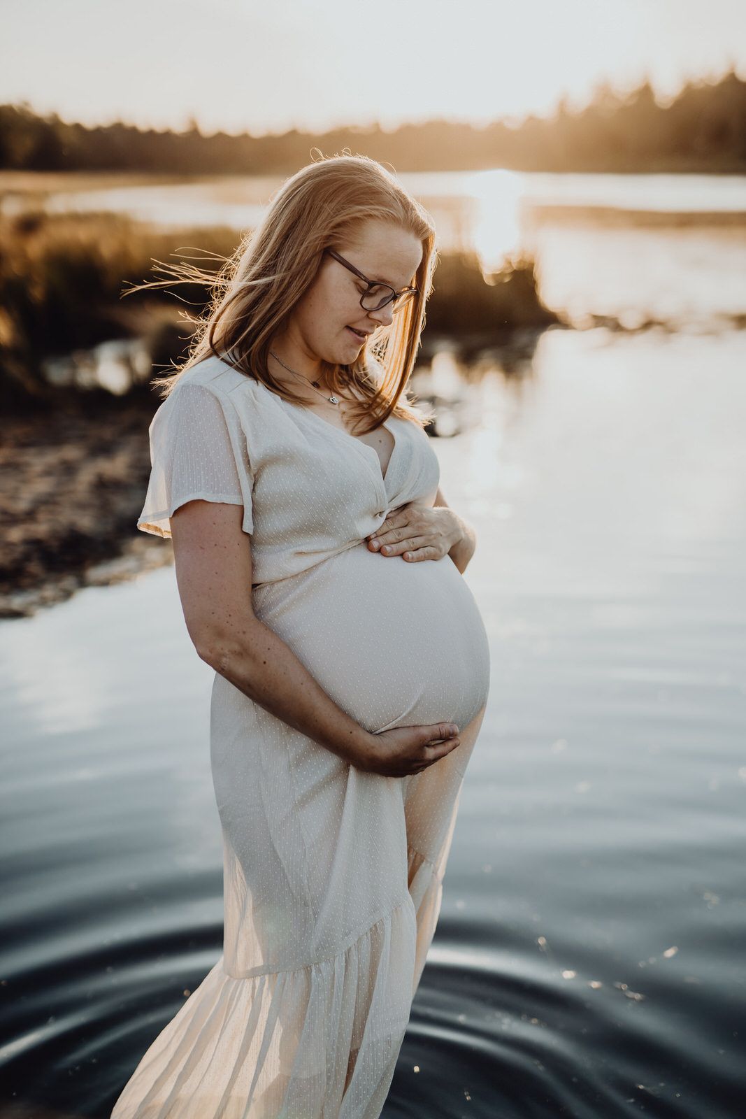 A maternity photo by the water at sunset showing a white dress and peaceful reflection.