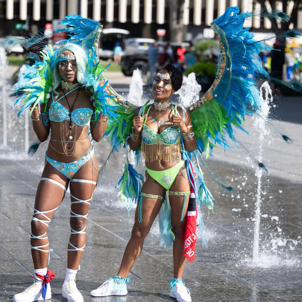 Carnival dancers in vibrant blue and green feathered costumes splash through fountain water during a festive parade.
