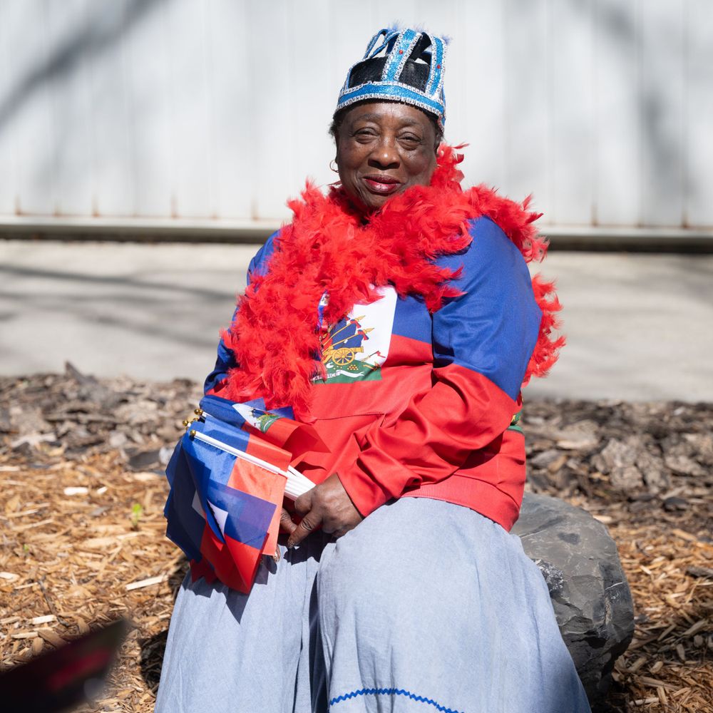 A person wearing a bright red feather boa and blue jacket sits on a concrete ledge in an outdoor setting.