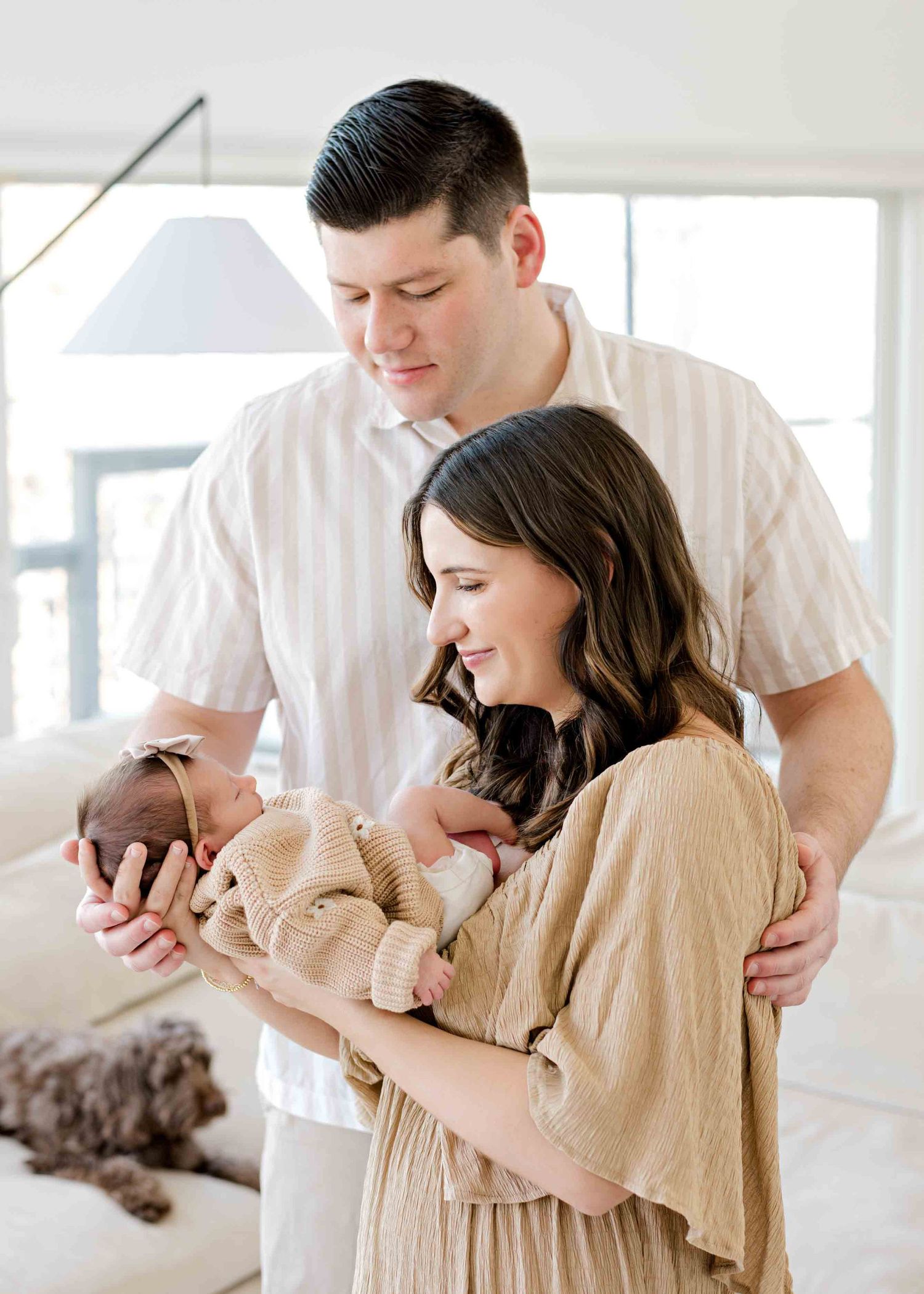 A tender family moment captured in a bright white living room with their infant and dog.