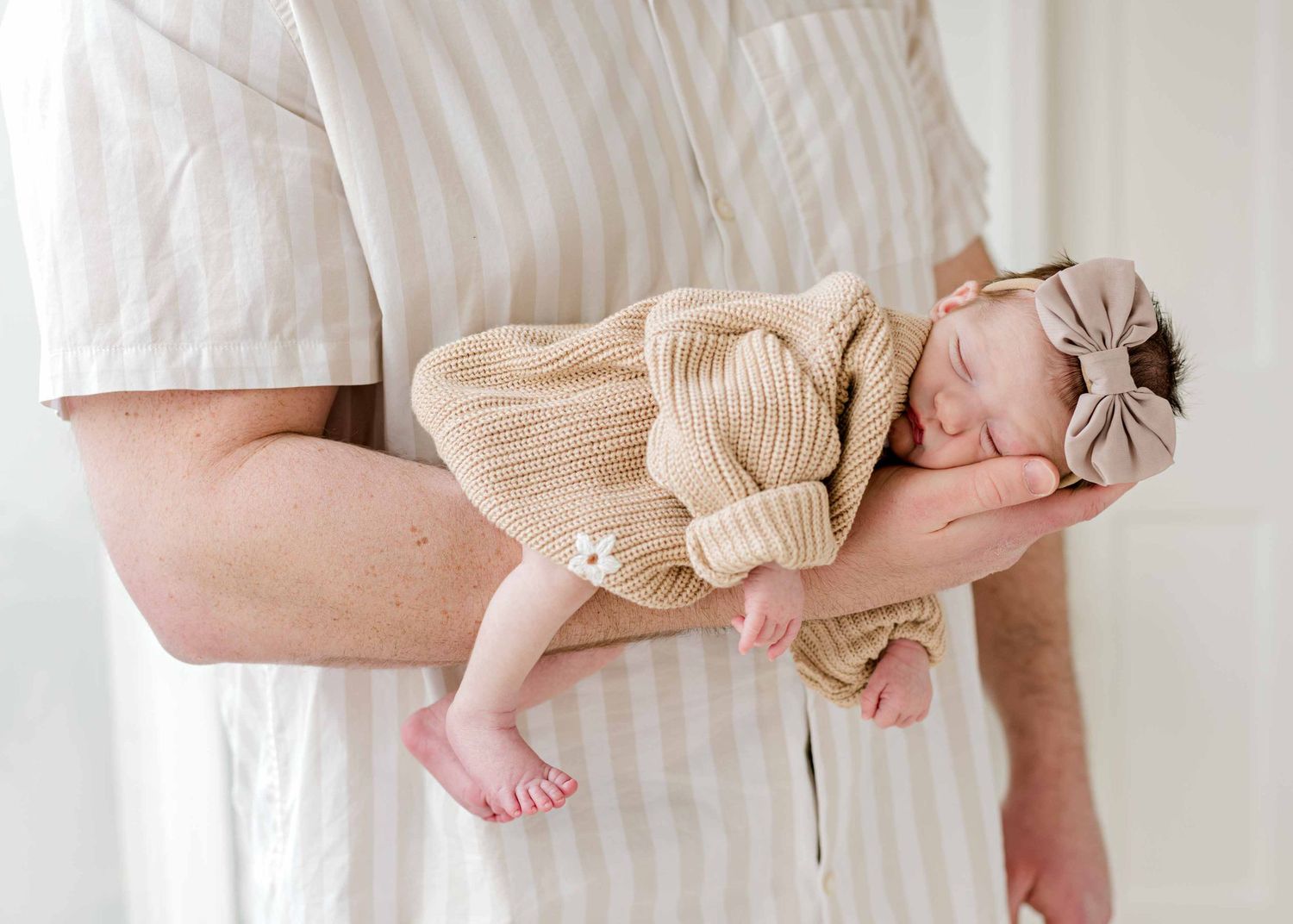 A sleeping newborn in beige knit clothing rests peacefully in supportive arms.