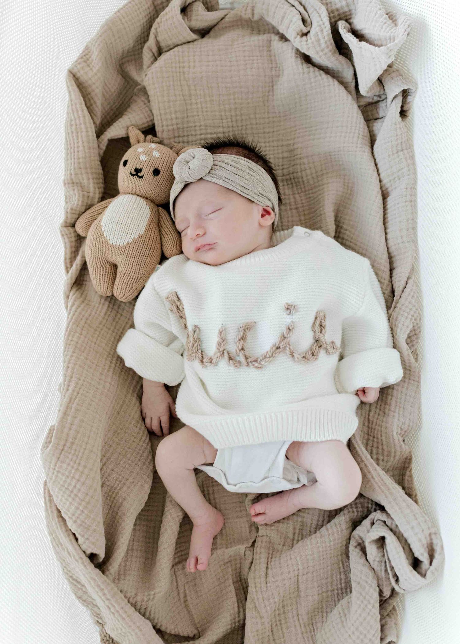Sleeping newborn in white knit sweater with stuffed bunny toy on beige blanket.