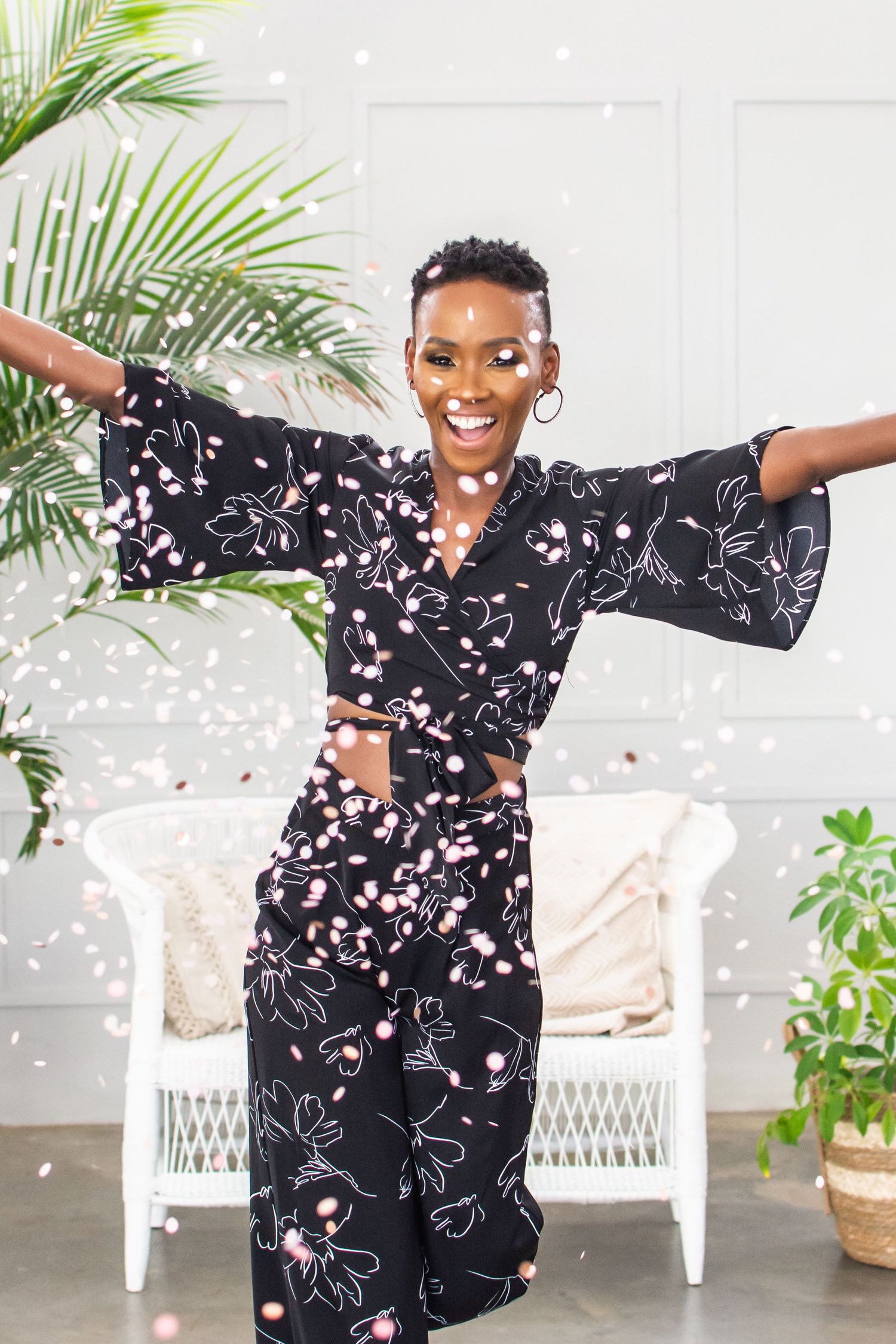 Joyful movement captured in a black floral dress against a white wicker chair and palm leaves background.