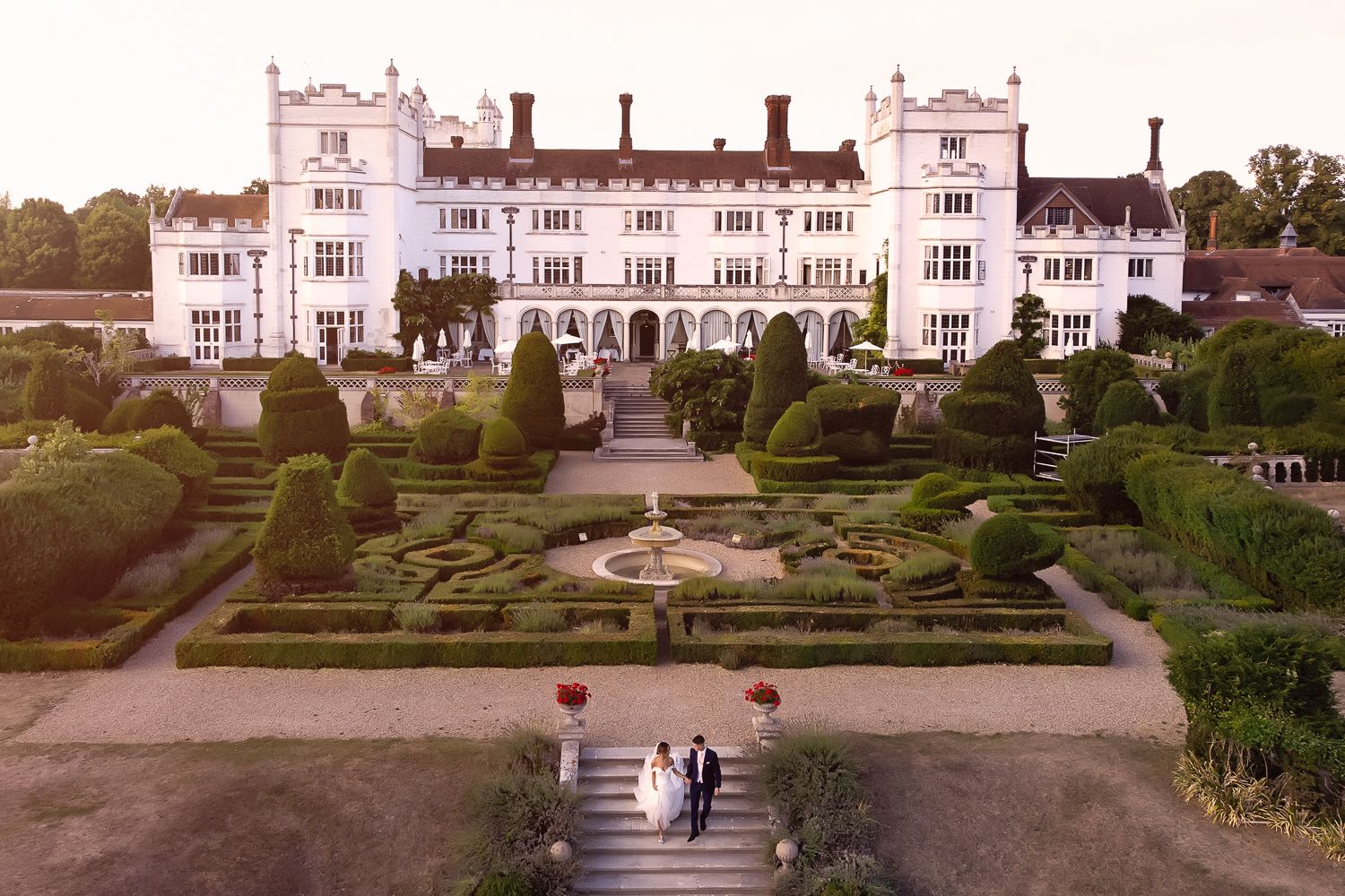Aerial view of a grand manor house with manicured gardens and a couple walking below.