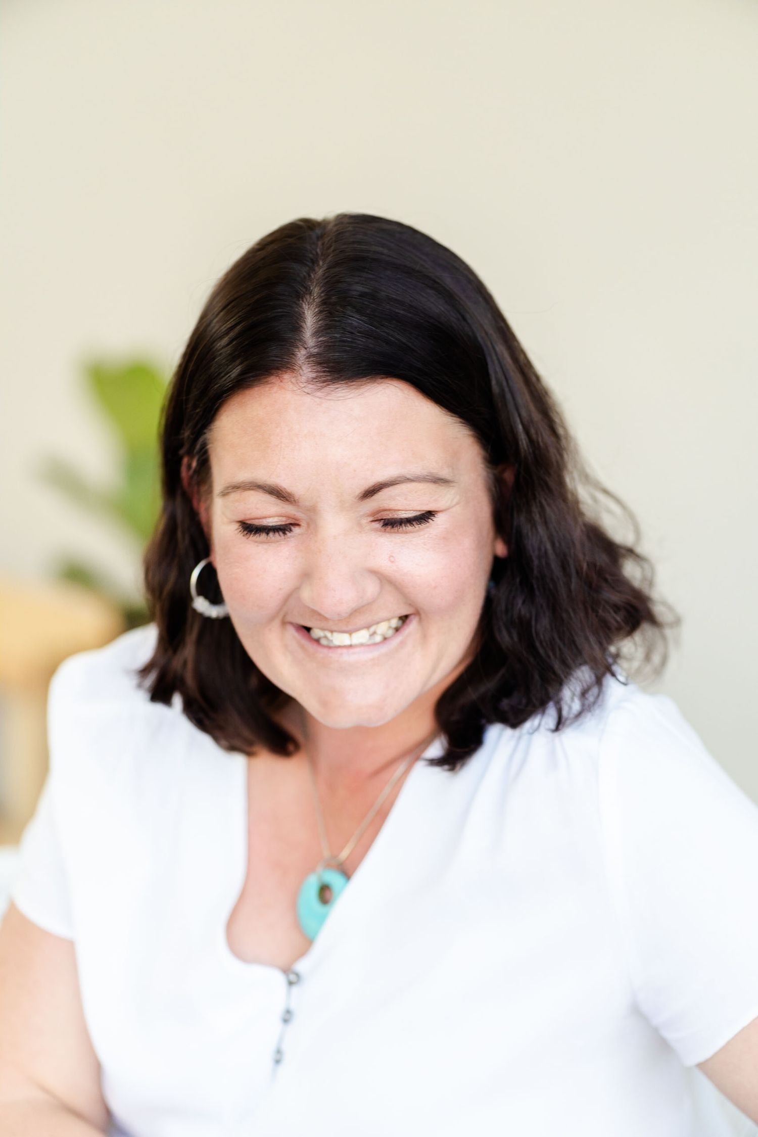 Person in white top smiles warmly while looking down with decorative pendant necklace.