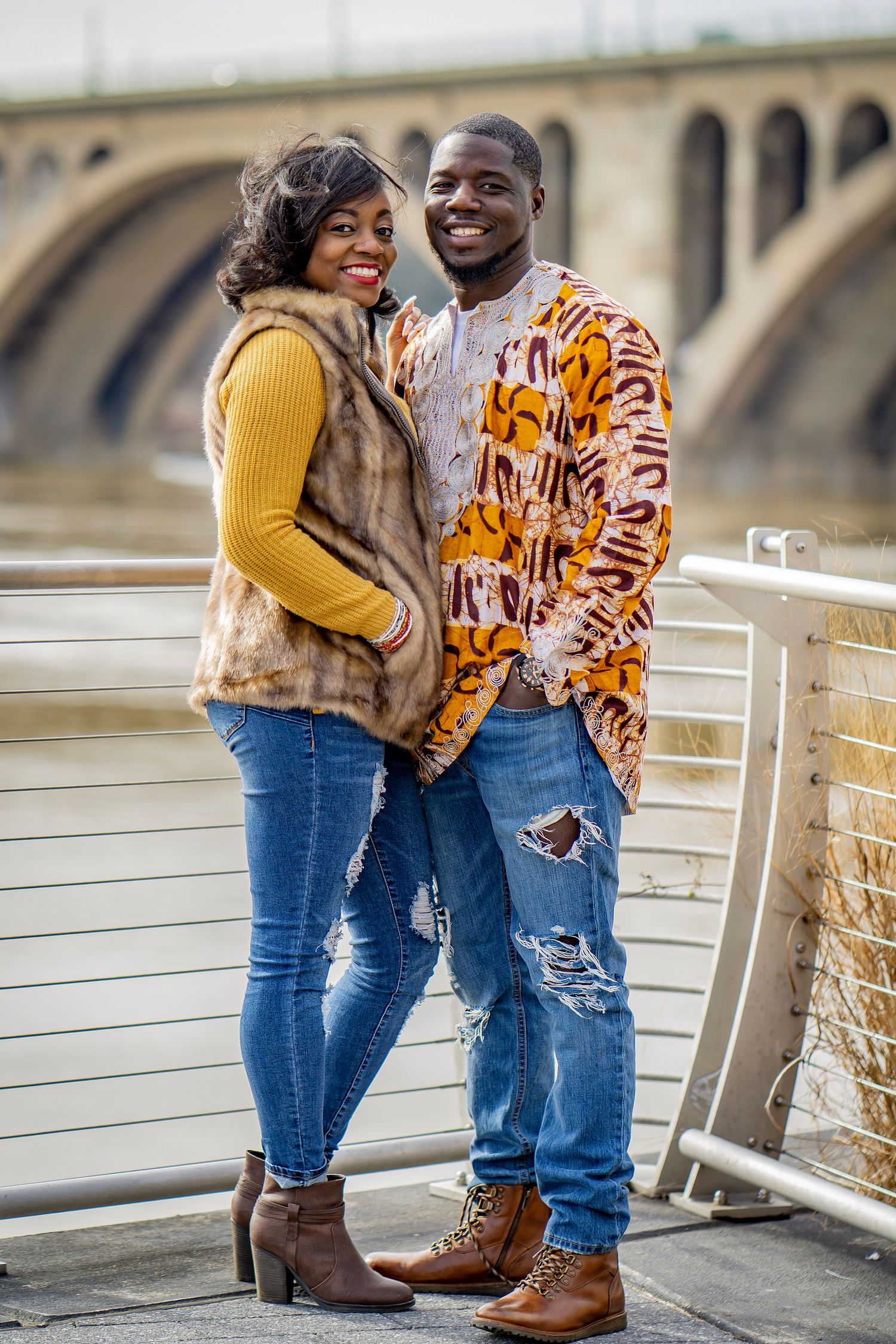 A couple in stylish winter outfits including a yellow sweater and patterned jacket pose together on a bridge.
