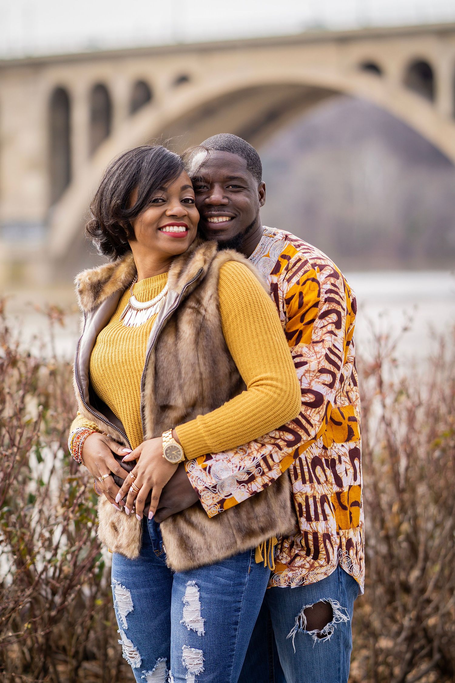 A couple in stylish winter wear embrace by a bridge with scenic architecture in the background.