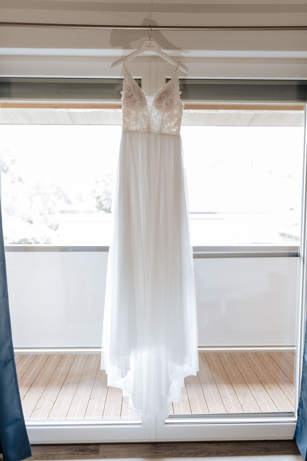 An elegant white lace wedding dress hanging on display in front of a bright window.