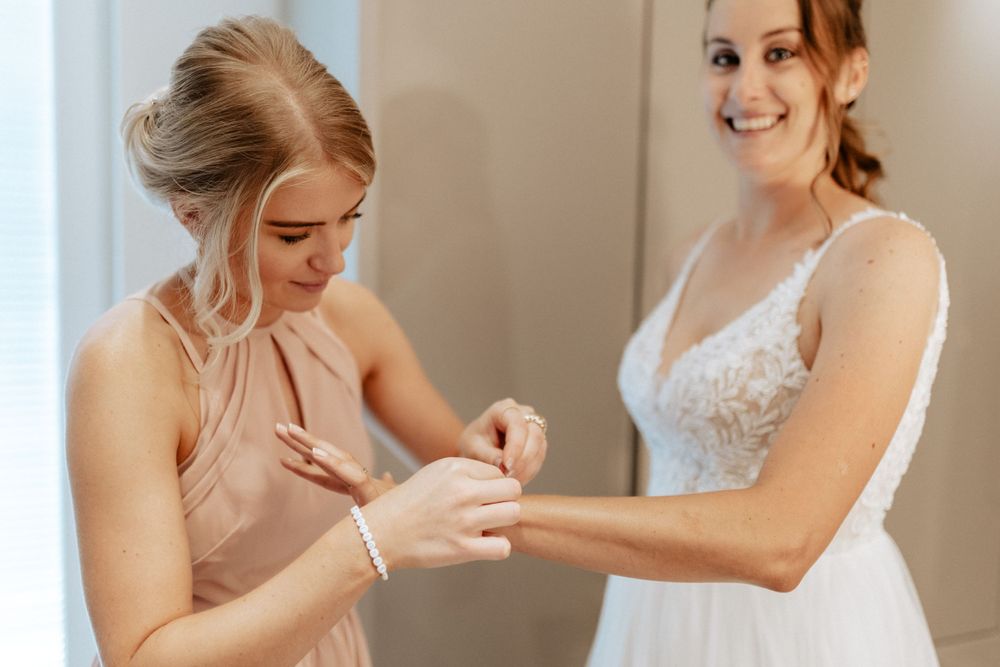 Two women in getting ready for a wedding as one helps adjust the other's bracelet.