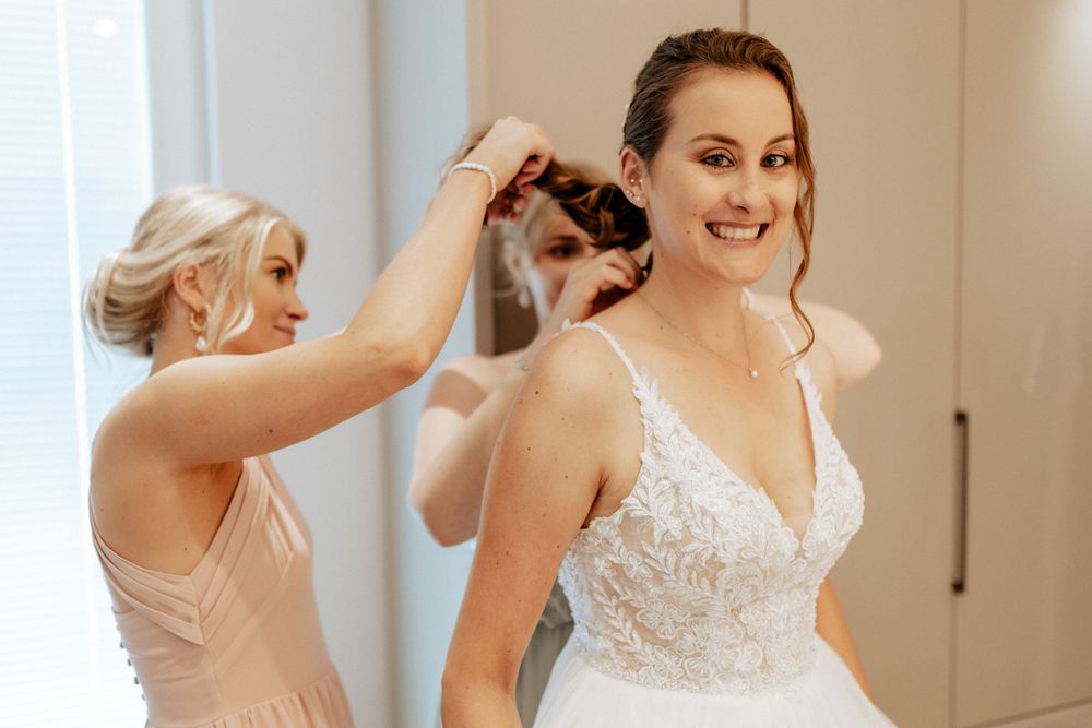A bride getting her hair styled while wearing a beautiful lace wedding dress.