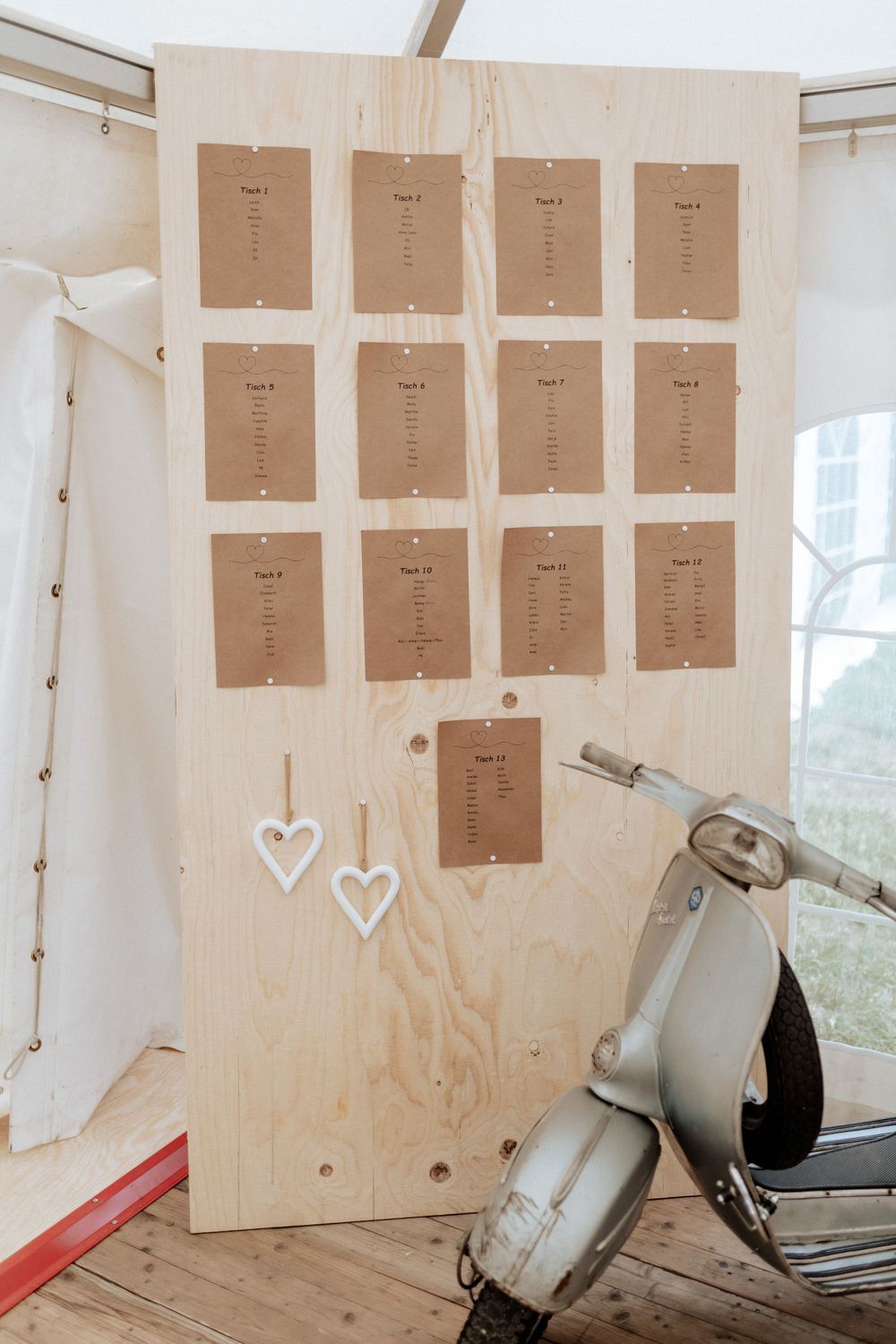 A rustic wooden seating chart display with brown paper cards and decorative hearts next to a vintage scooter.