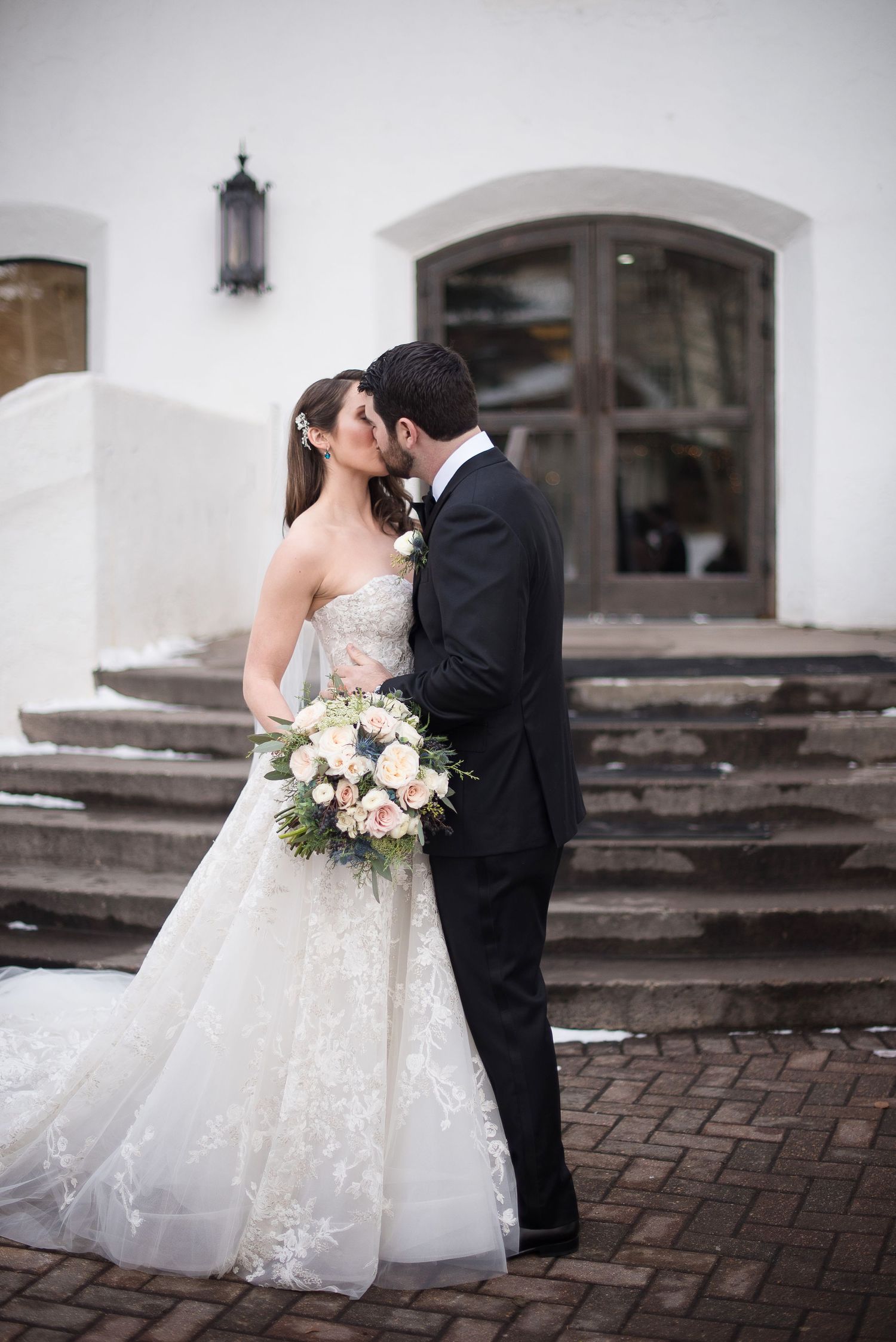 bride in a strapless lace monique lhullier gown and groom in classic black tux kiss in front of church steps in Vail, CO